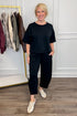 Front view of woman wearing Swagger Boutique’s Black Round Neck Short Sleeve Top with matching barrel pants with clothes hanging in the background.