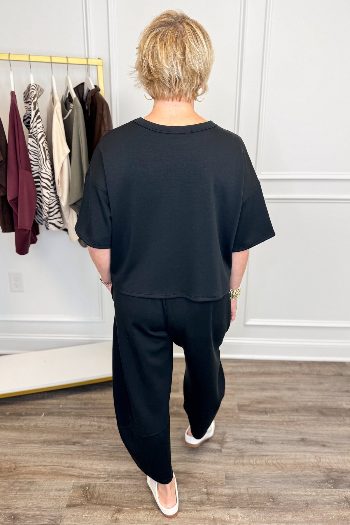 Back view of woman wearing Swagger Boutique’s Black Round Neck Short Sleeve Top with matching barrel pants with clothes hanging in the background.