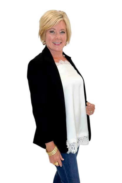 Side view of woman wearing Swagger Boutique’s Black Blazer and Ivory Satin Lace Trim Tank Top by Entro with jeans against white studio background.