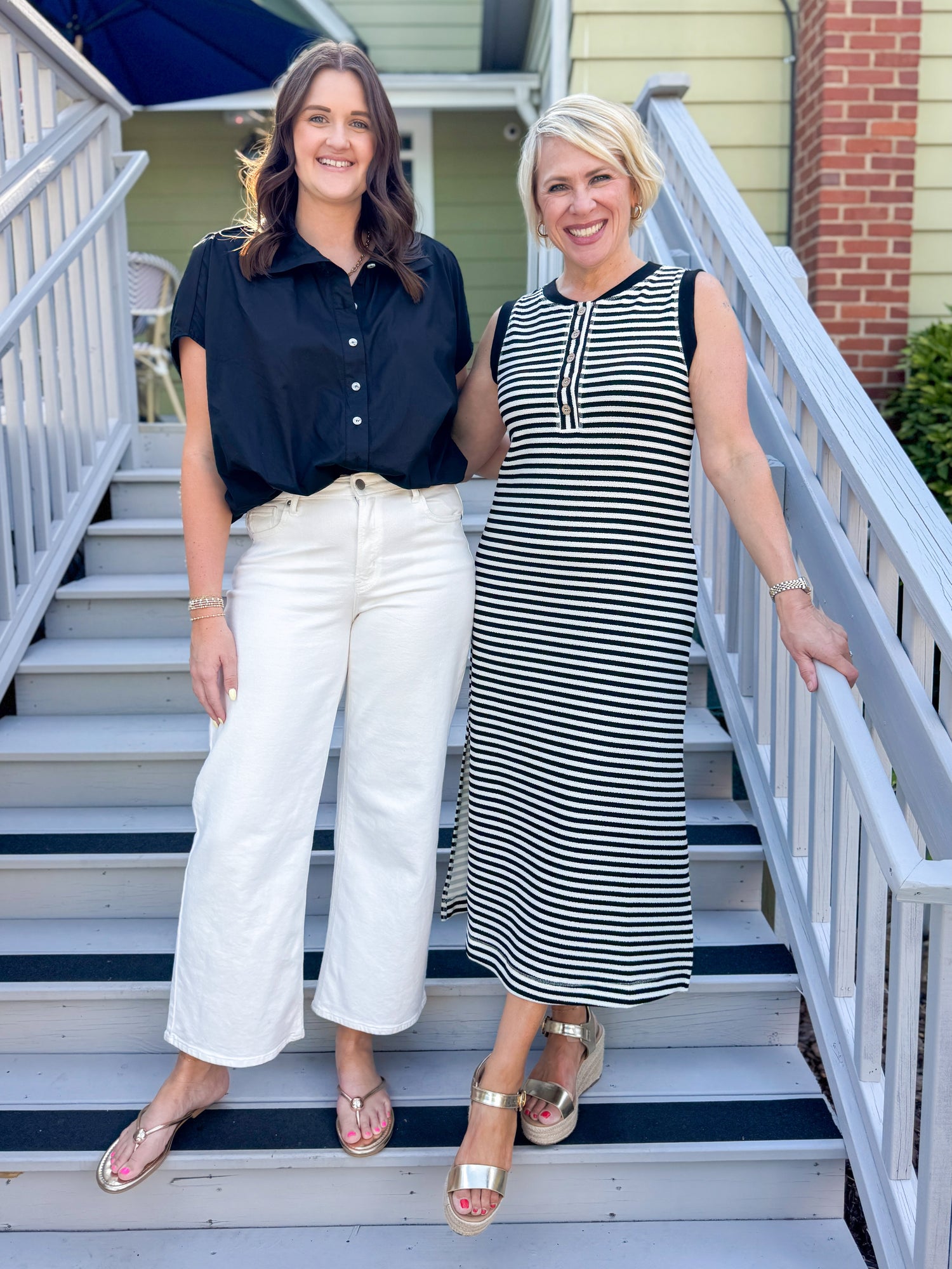 Front view of woman wearing Swagger Boutique’s Black Collared Bubble Hem Top with jeans and woman in striped dress standing outside a house.