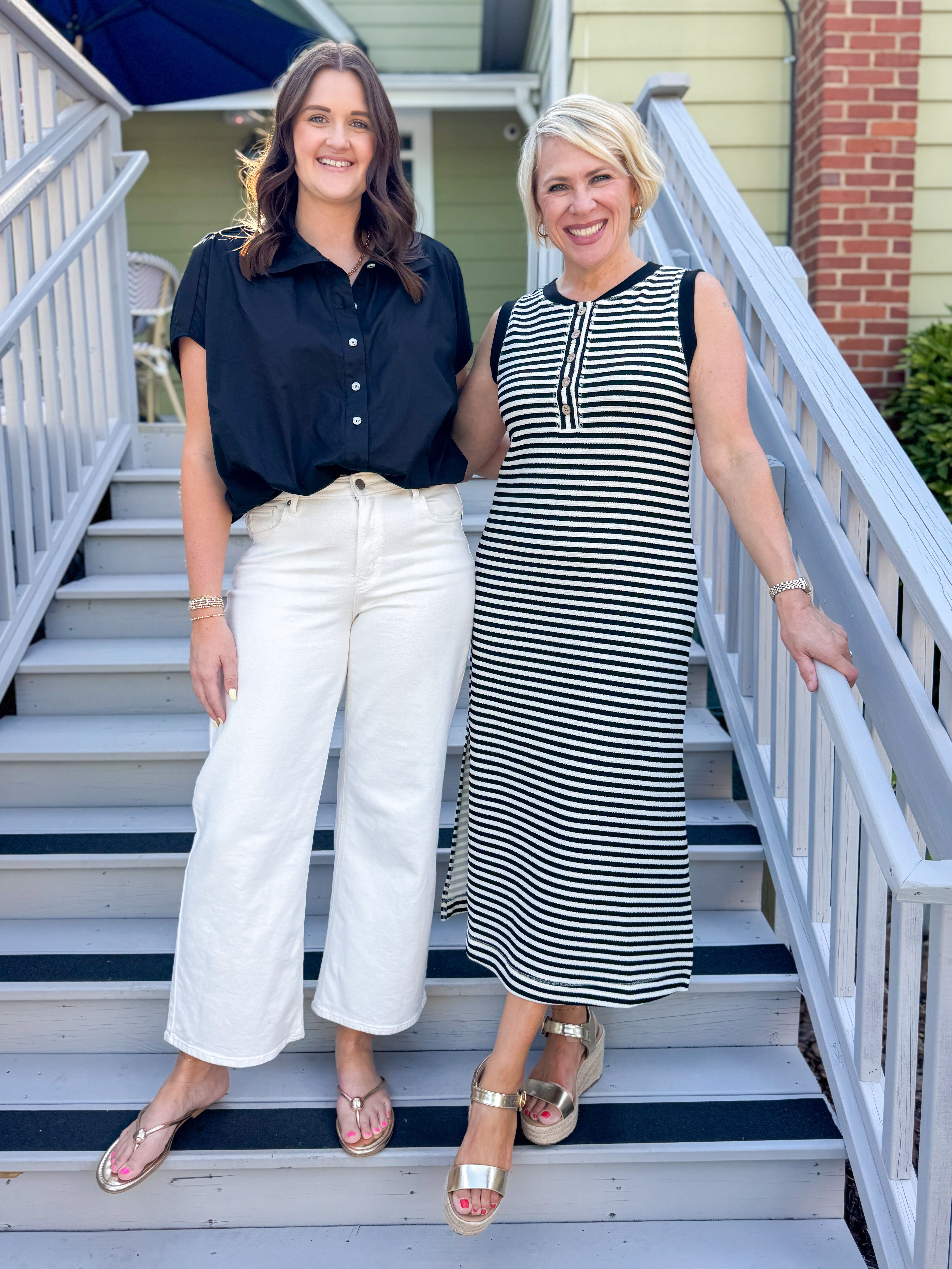 Front view of woman wearing Swagger Boutique’s Black Collared Bubble Hem Top with jeans and woman in striped dress standing outside a house.