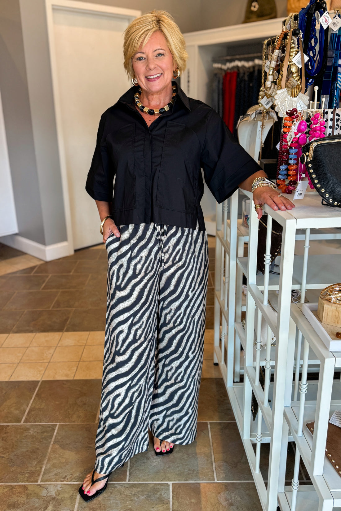 Front view of woman wearing Swagger Boutique’s Black Collared Short Sleeve Top with Zebra Wide Leg Pants by Entro against standing at the store.