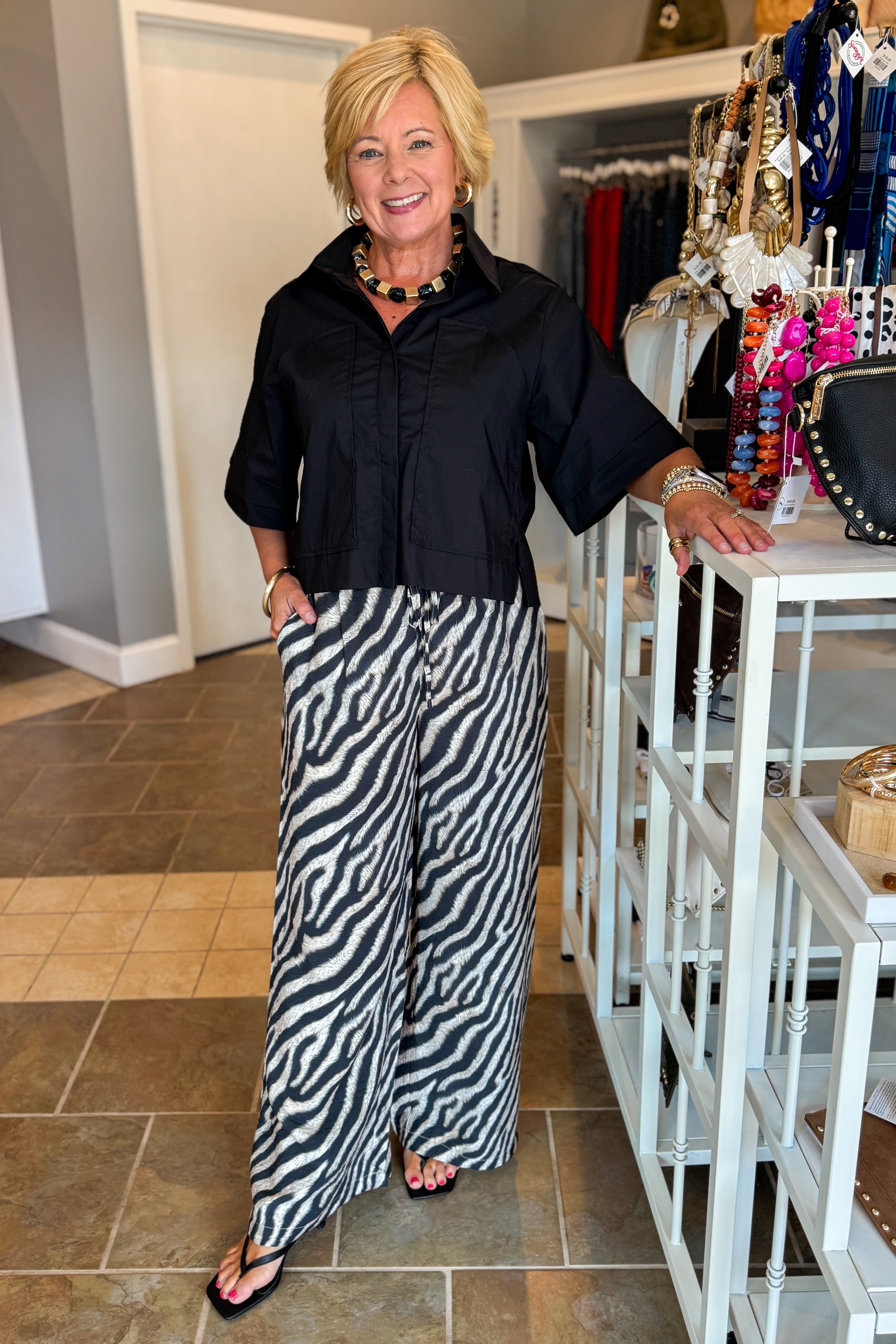 Front view of woman wearing Swagger Boutique’s Black Collared Short Sleeve Top with Zebra Wide Leg Pants by Entro against standing at the store.