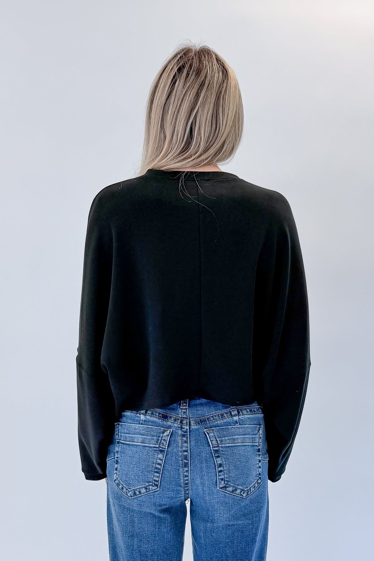 Back view of woman wearing Swagger Boutique’s black cropped sweatshirt with jeans against white studio background.