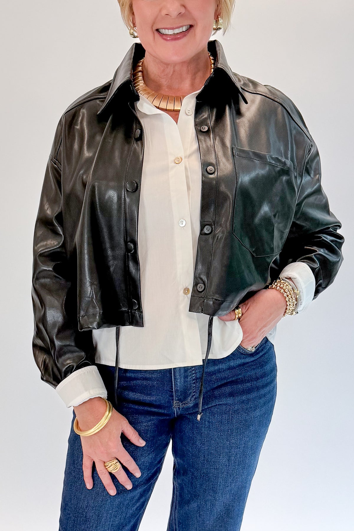 Front detail view of woman wearing Swagger Boutique’s Black Faux Leather Jacket With Layered Shirt with jeans against white studio background.