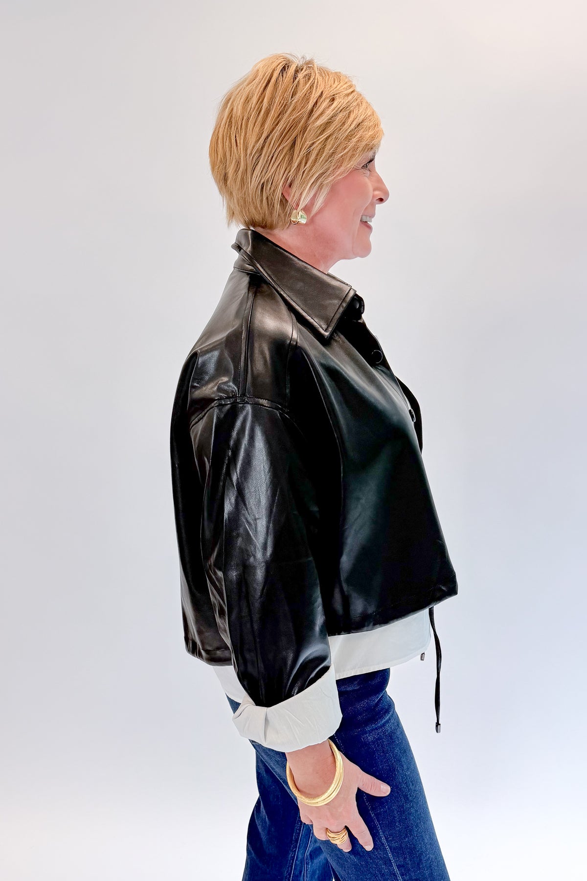 Side view of woman wearing Swagger Boutique’s Black Faux Leather Jacket With Layered Shirt with jeans against white studio background.