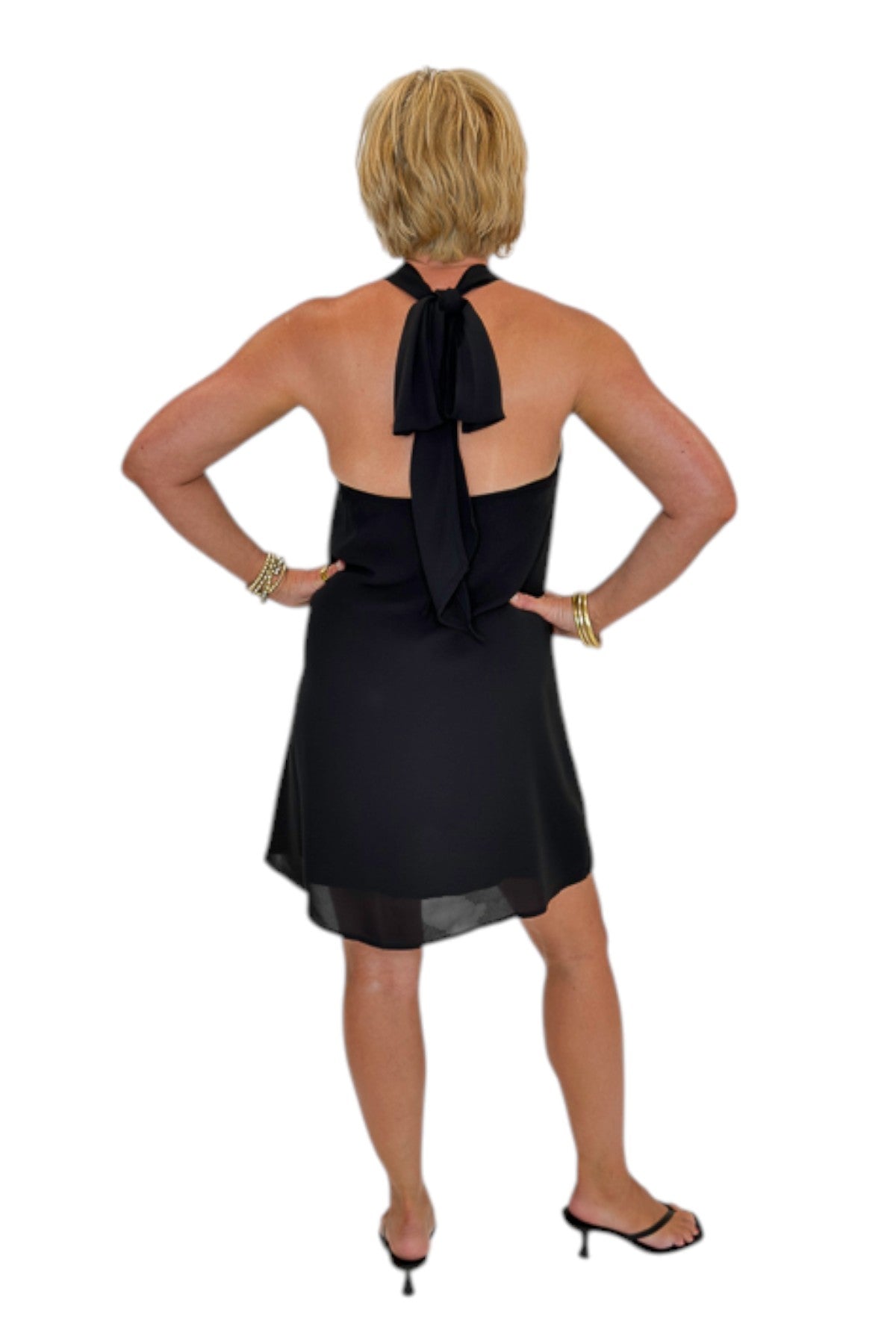 Back view of woman wearing Swagger Boutique’s Black Halter Neck Mini Dress with heels against white studio background.