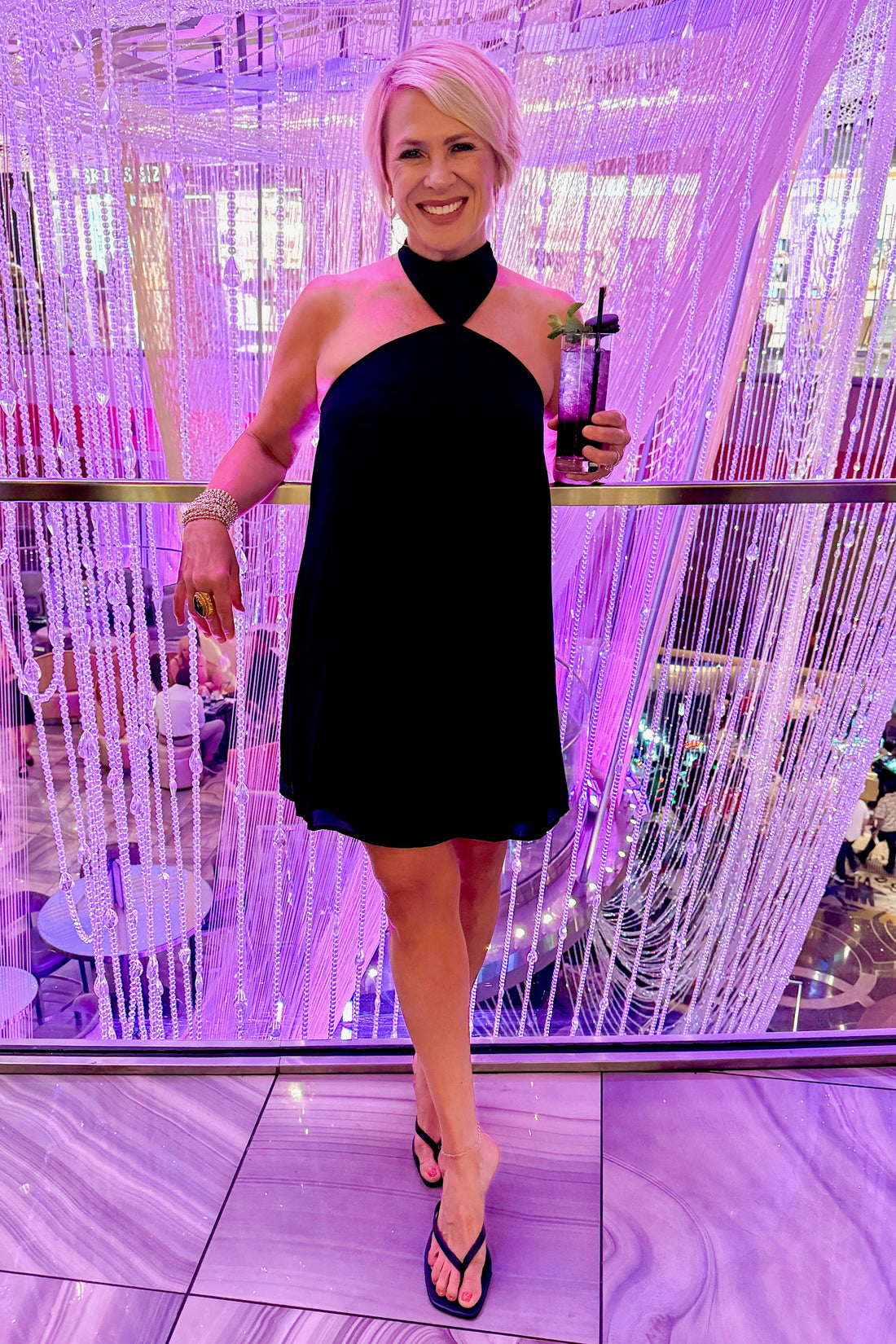 Front view of woman wearing Swagger Boutique’s Black Halter Neck Mini Dress with heels at a fancy restaurant. 