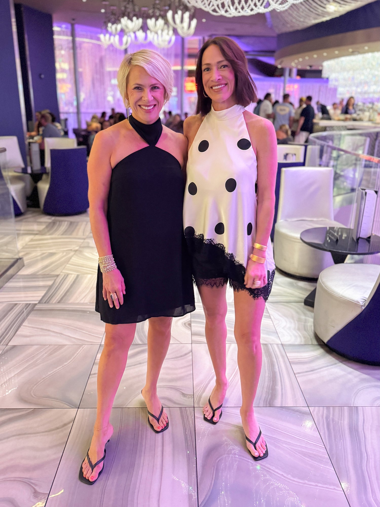 Front view of women wearing Swagger Boutique’s Black Halter Neck Mini Dress with heels and polka dot halter top at a fancy restaurant. 