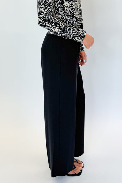 Side detail view of woman wearing Swagger Boutique’s Black High Waisted Trousers by Skies Are Blue &amp; Zebra Mock Neck Long Sleeve Top by Entro against white studio background.
