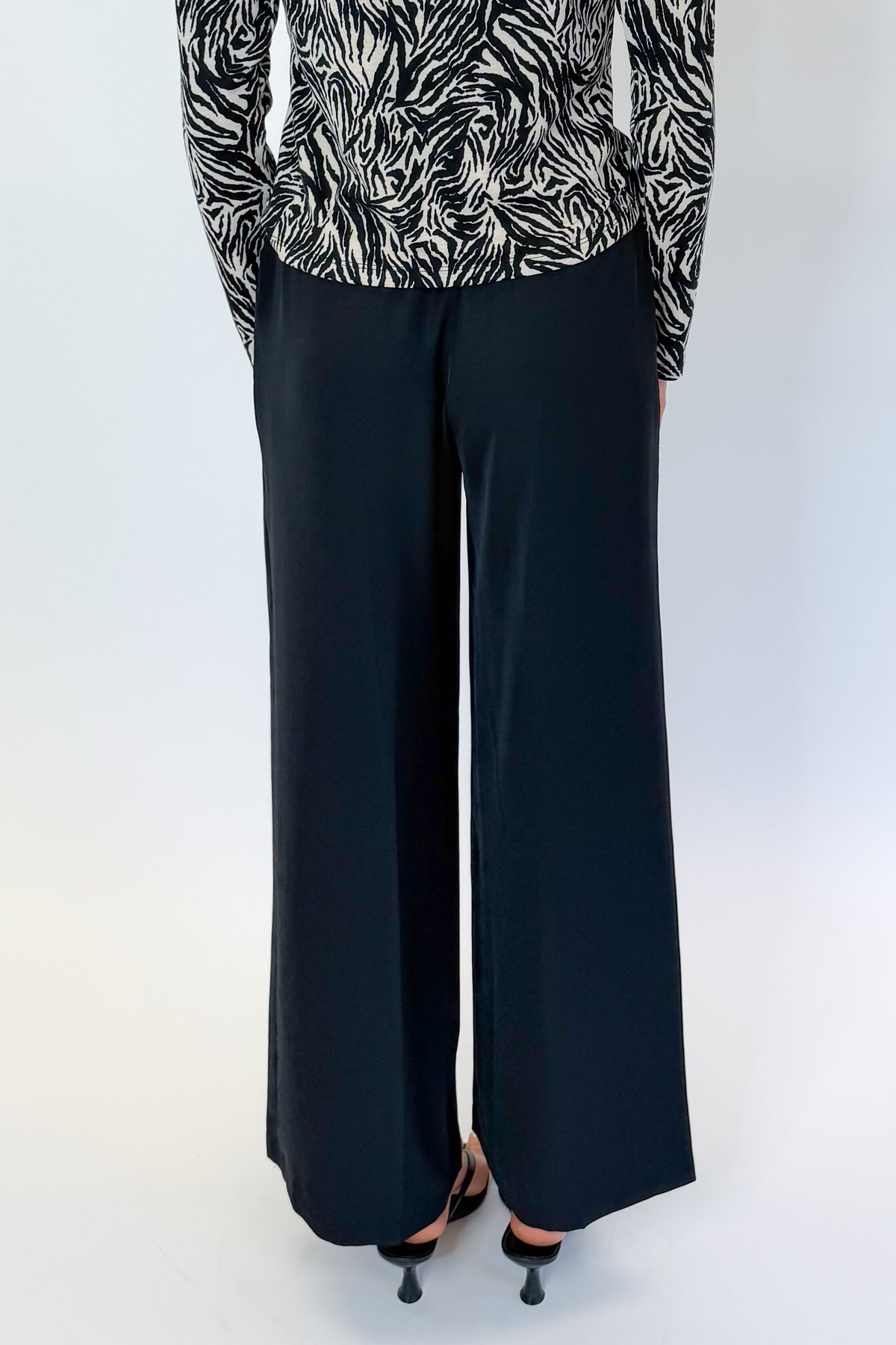 Back view of woman wearing Swagger Boutique’s Black High Waisted Trousers by Skies Are Blue &amp; Zebra Mock Neck Long Sleeve Top by Entro against white studio background.