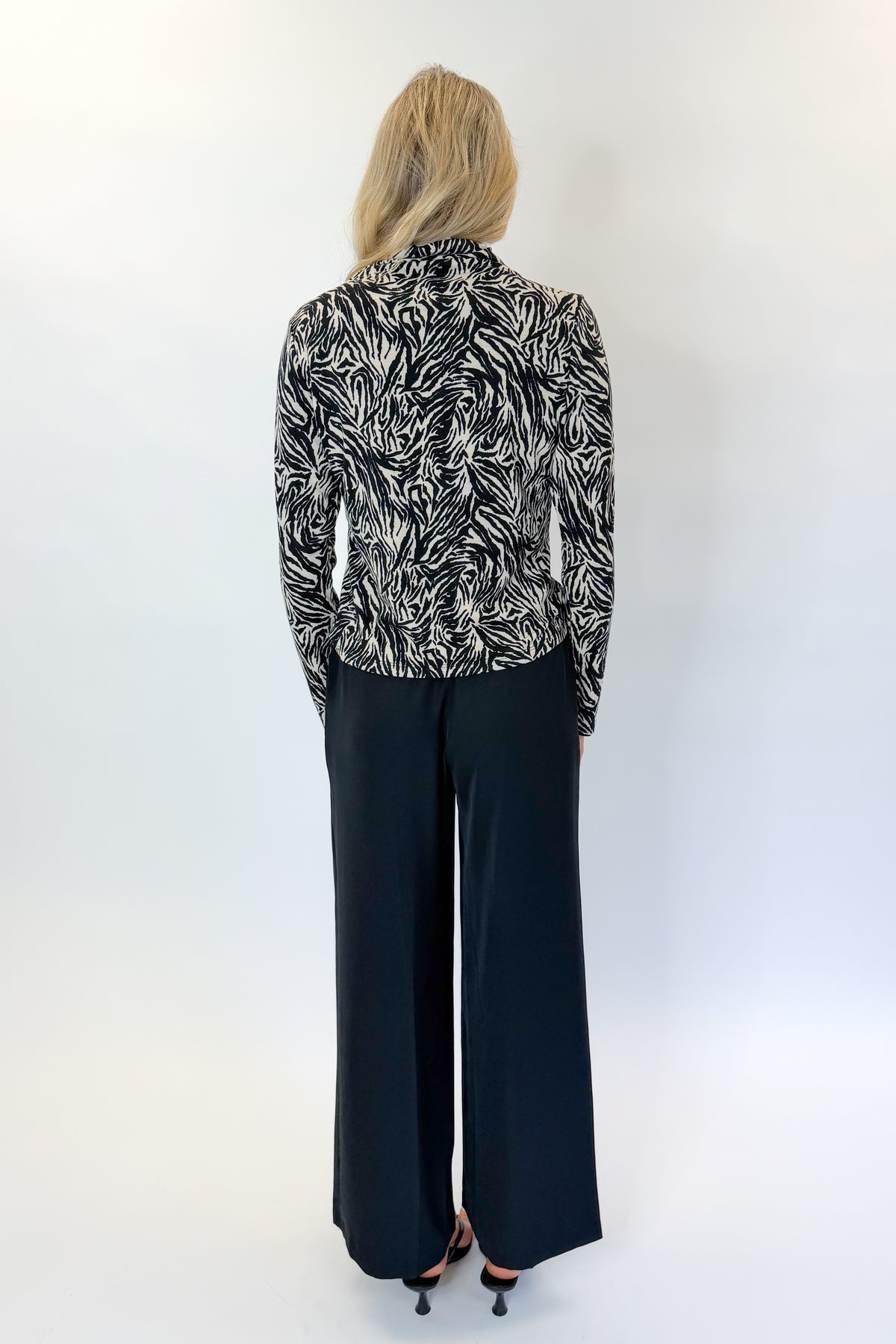 Back view of woman wearing Swagger Boutique’s Black High Waisted Trousers by Skies Are Blue &amp; Zebra Mock Neck Long Sleeve Top by Entro against white studio background.