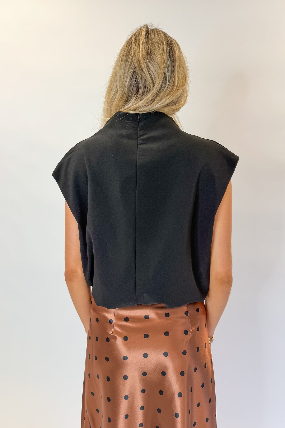 Back view of woman wearing Swagger Boutique’s Camel Polka Dot Satin Skirt with Black Mock Neck Bubble Hem Tank against white studio background.
