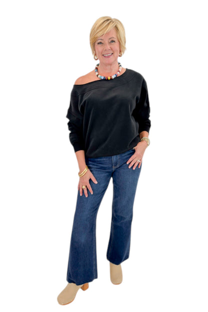 Front view of woman wearing Swagger Boutique’s Black Off The Shoulder Sweatshirt with jeans against white studio background.