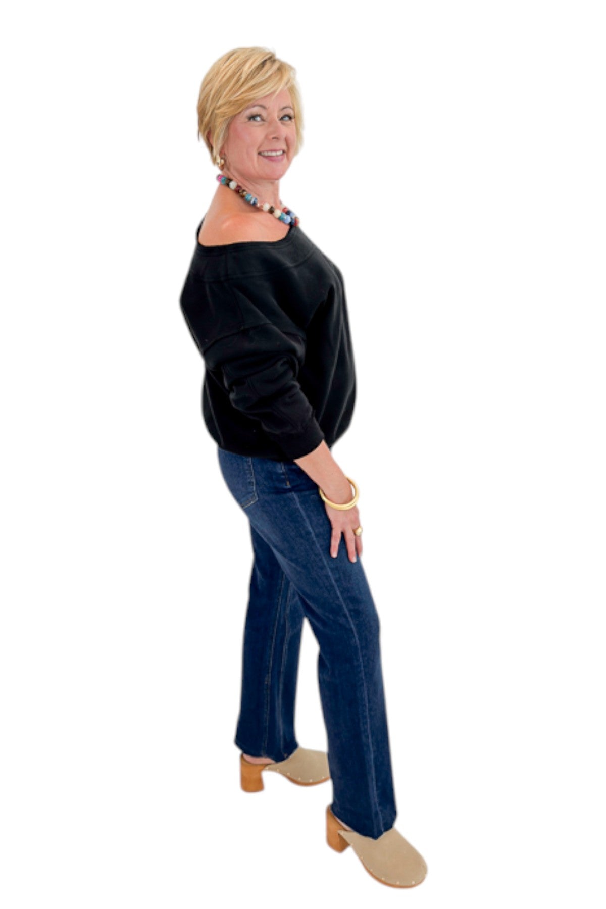 Side view of woman wearing Swagger Boutique’s Black Off The Shoulder Sweatshirt with jeans against white studio background.