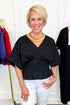 Front view of woman wearing Swagger Boutique’s Black Pleat Detail Dolman Sleeve Top with jeans with clothing hanging in the background.