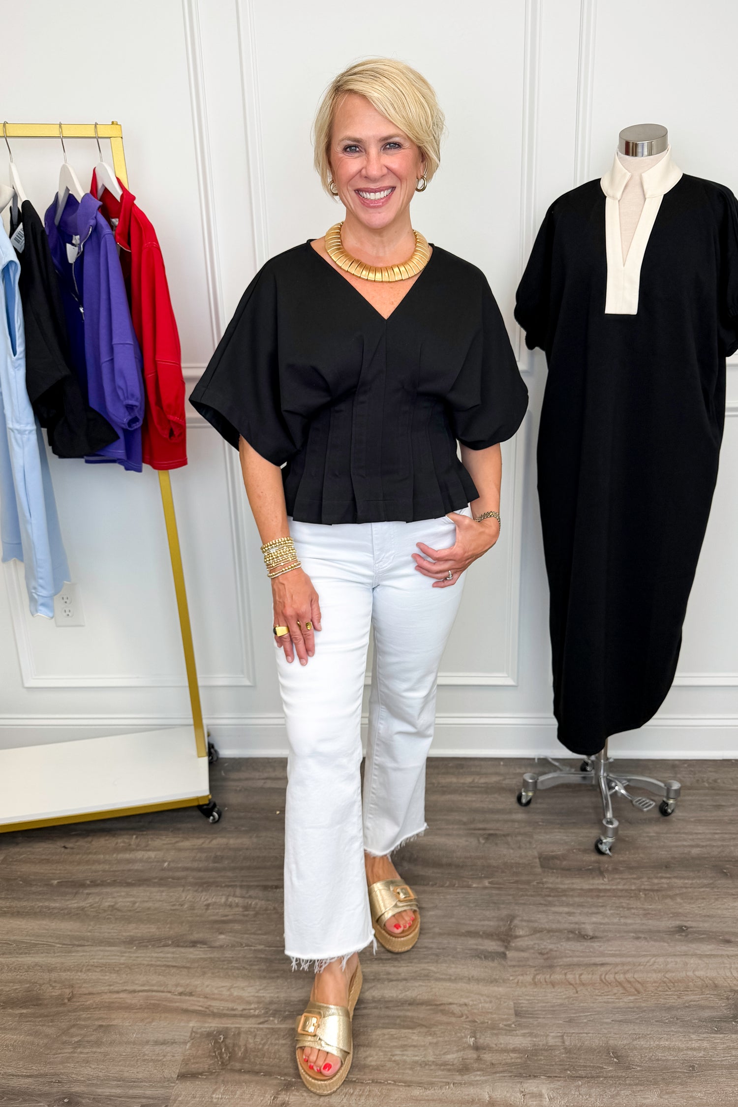 Front view of woman wearing Swagger Boutique’s Black Pleat Detail Dolman Sleeve Top with jeans with clothing hanging in the background.