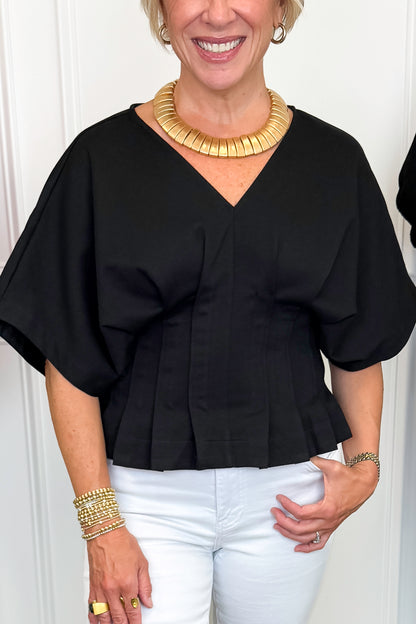 Front detail view of woman wearing Swagger Boutique’s Black Pleat Detail Dolman Sleeve Top with jeans with clothing hanging in the background.