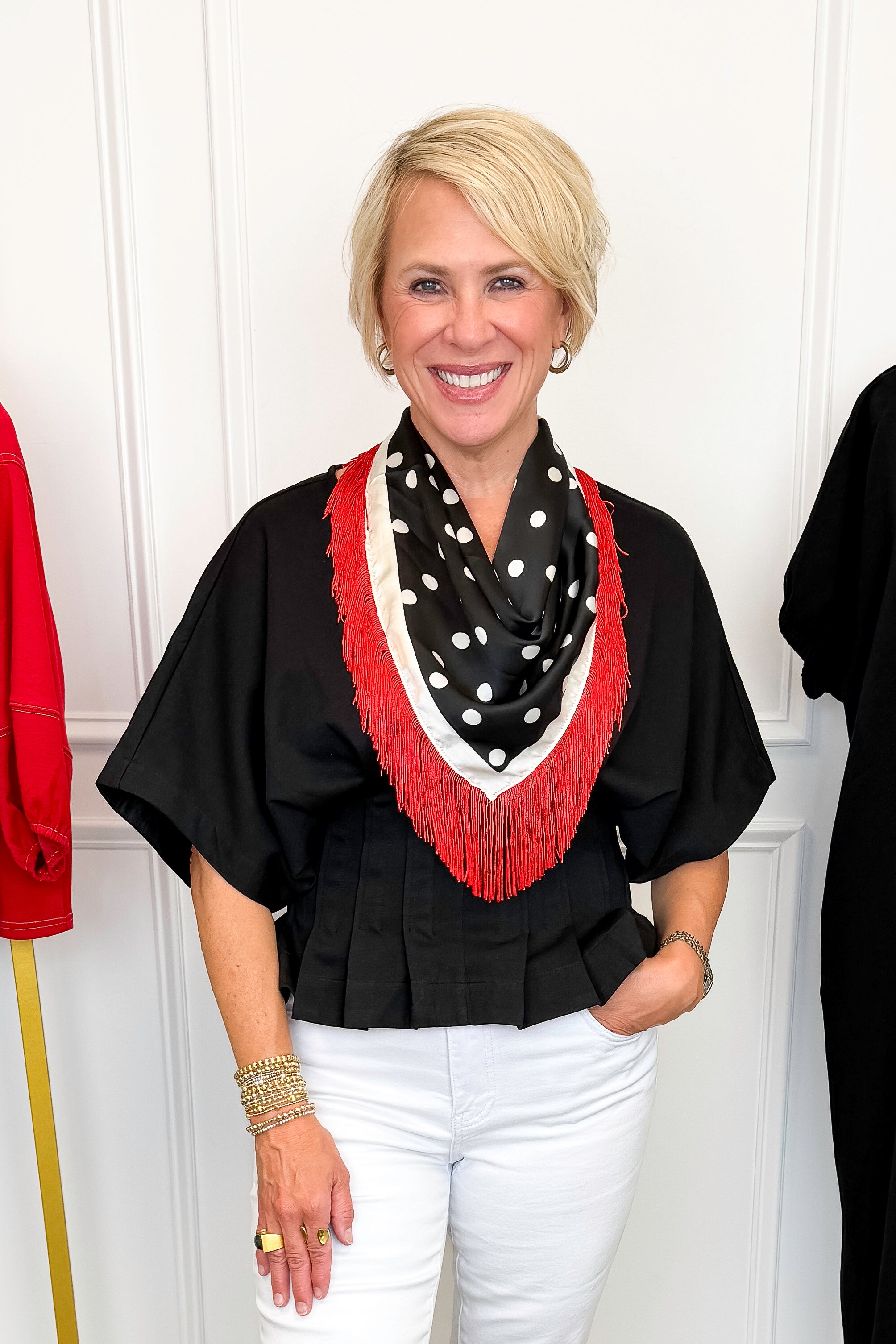 Front view of woman wearing Swagger Boutique’s Black Pleat Detail Dolman Sleeve Top with jeans with clothing hanging in the background.