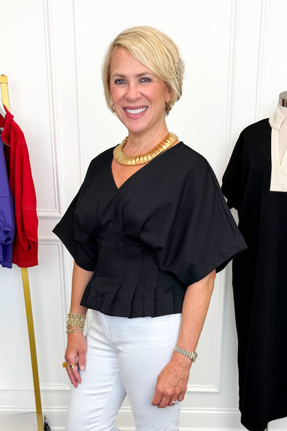 Side view of woman wearing Swagger Boutique’s Black Pleat Detail Dolman Sleeve Top with jeans with clothing hanging in the background.