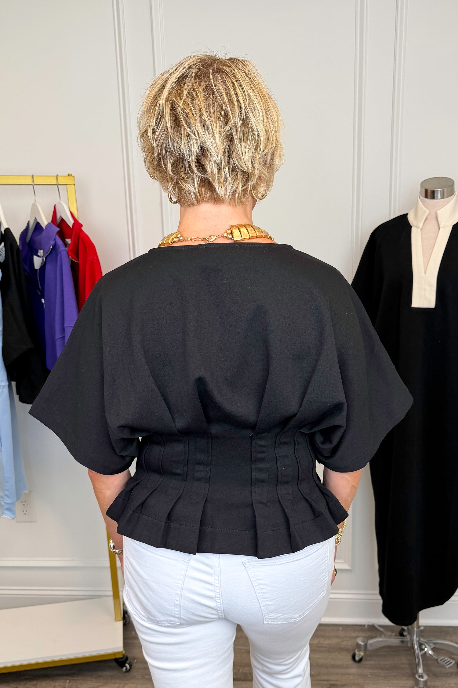 Back view of woman wearing Swagger Boutique’s Black Pleat Detail Dolman Sleeve Top with jeans with clothing hanging in the background.