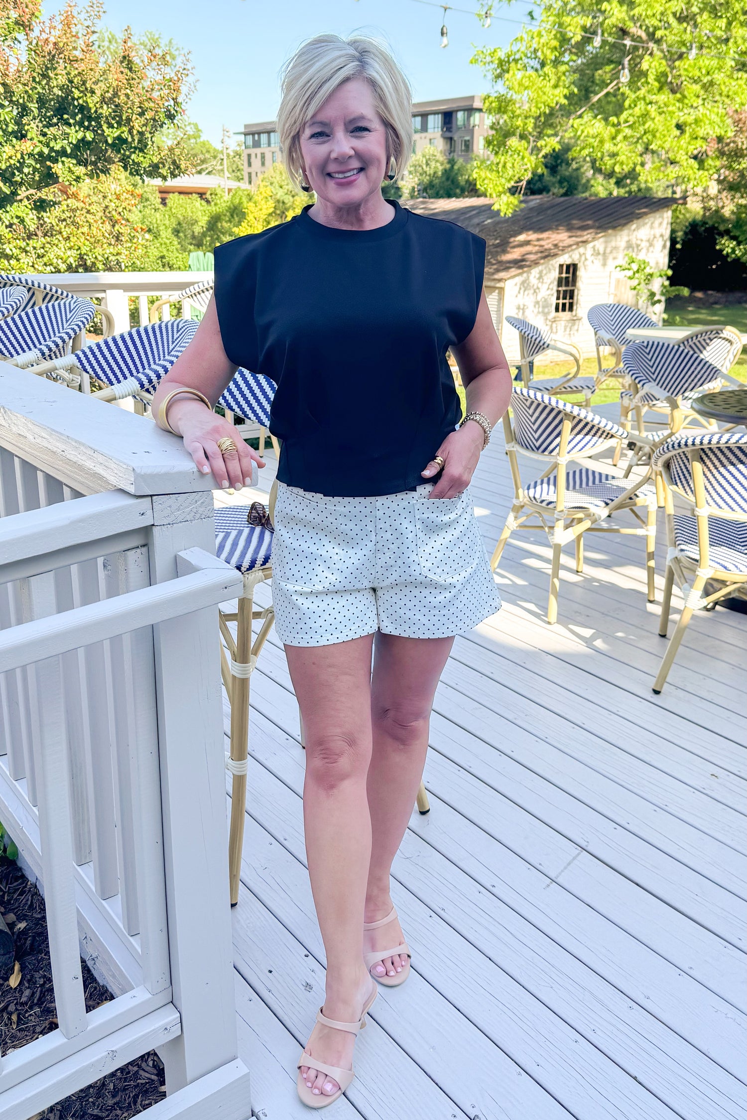 Front view of woman wearing Swagger Boutique’s Black Pleat Detail Tank with cream and black polka dot shorts standing outside at a restaurant.