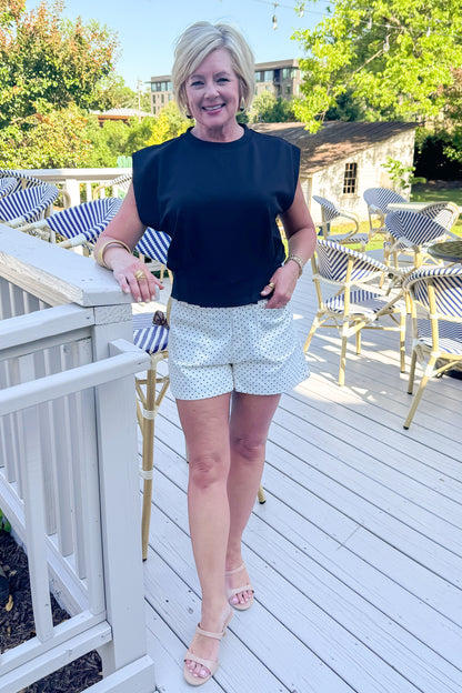 Front view of woman wearing Swagger Boutique’s Black Pleat Detail Tank with cream and black polka dot shorts standing outside at a restaurant.