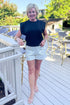 Front view of woman wearing Swagger Boutique’s Black Pleat Detail Tank with cream and black polka dot shorts standing outside at a restaurant.