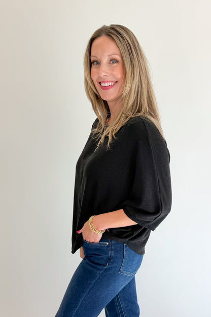 Side view of woman wearing Swagger Boutique’s Black Ribbed Dolman Sleeve Top with jeans against white studio background.
