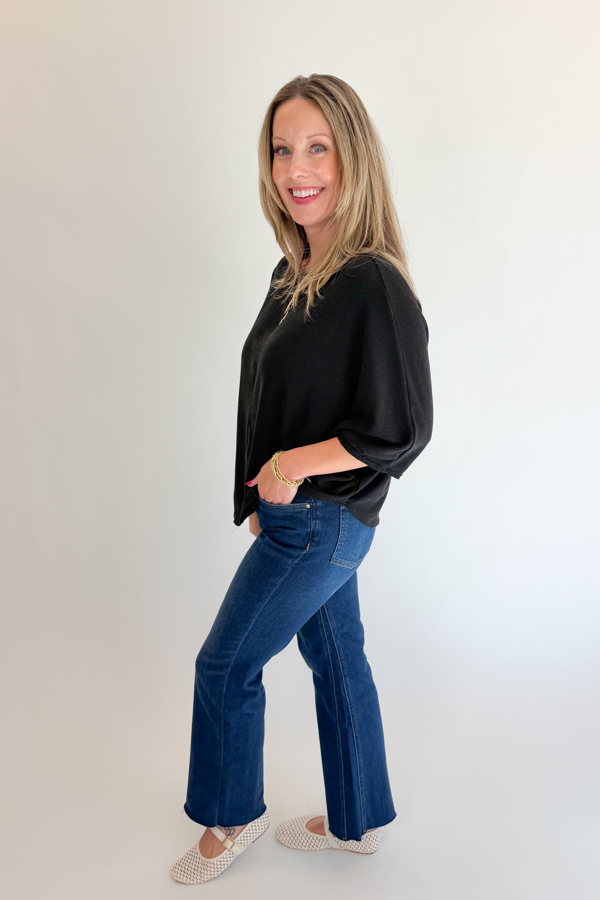 Side view of woman wearing Swagger Boutique’s Black Ribbed Dolman Sleeve Top with jeans against white studio background.