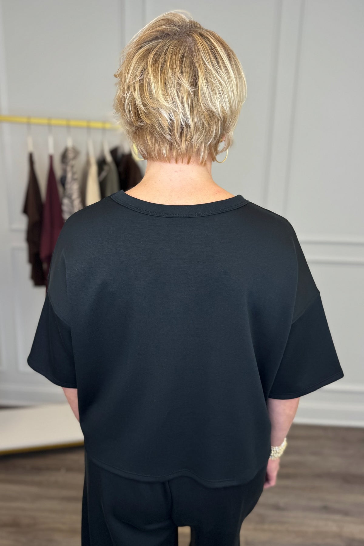 Back view of woman wearing Swagger Boutique’s Black Round Neck Short Sleeve Top with matching barrel pants with clothes hanging in the background.