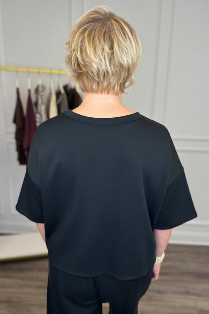 Back view of woman wearing Swagger Boutique’s Black Round Neck Short Sleeve Top with matching barrel pants with clothes hanging in the background.