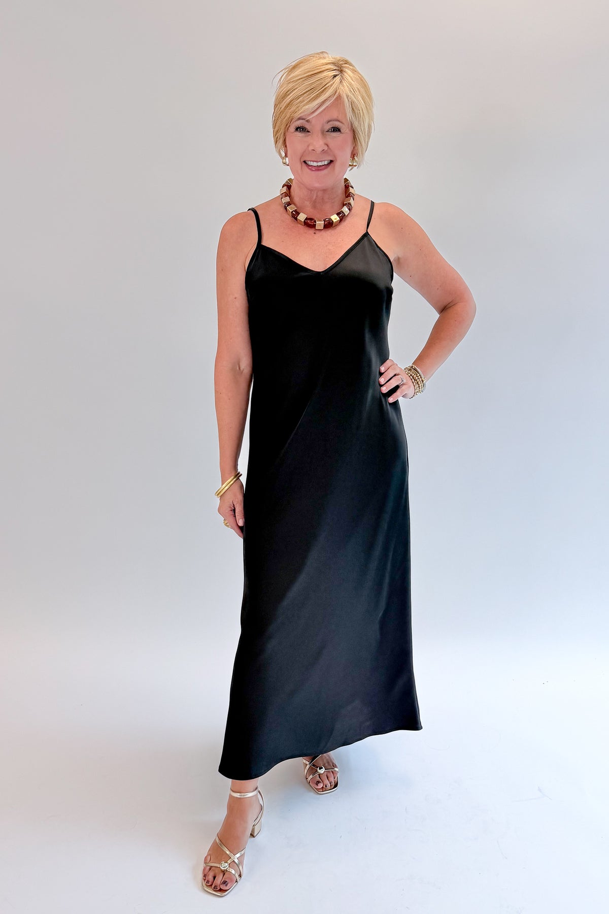 Front view of woman wearing Swagger Boutique’s Black Satin Dress with heels against white studio background.