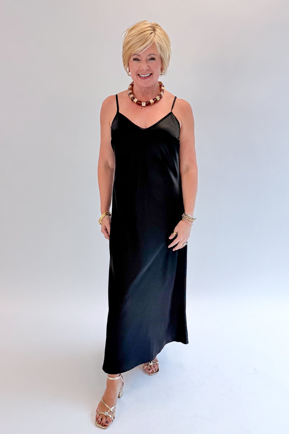 Front view of woman wearing Swagger Boutique’s Black Satin Dress with heels against white studio background.