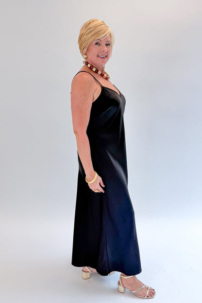 Side view of woman wearing Swagger Boutique’s Black Satin Dress with heels against white studio background.