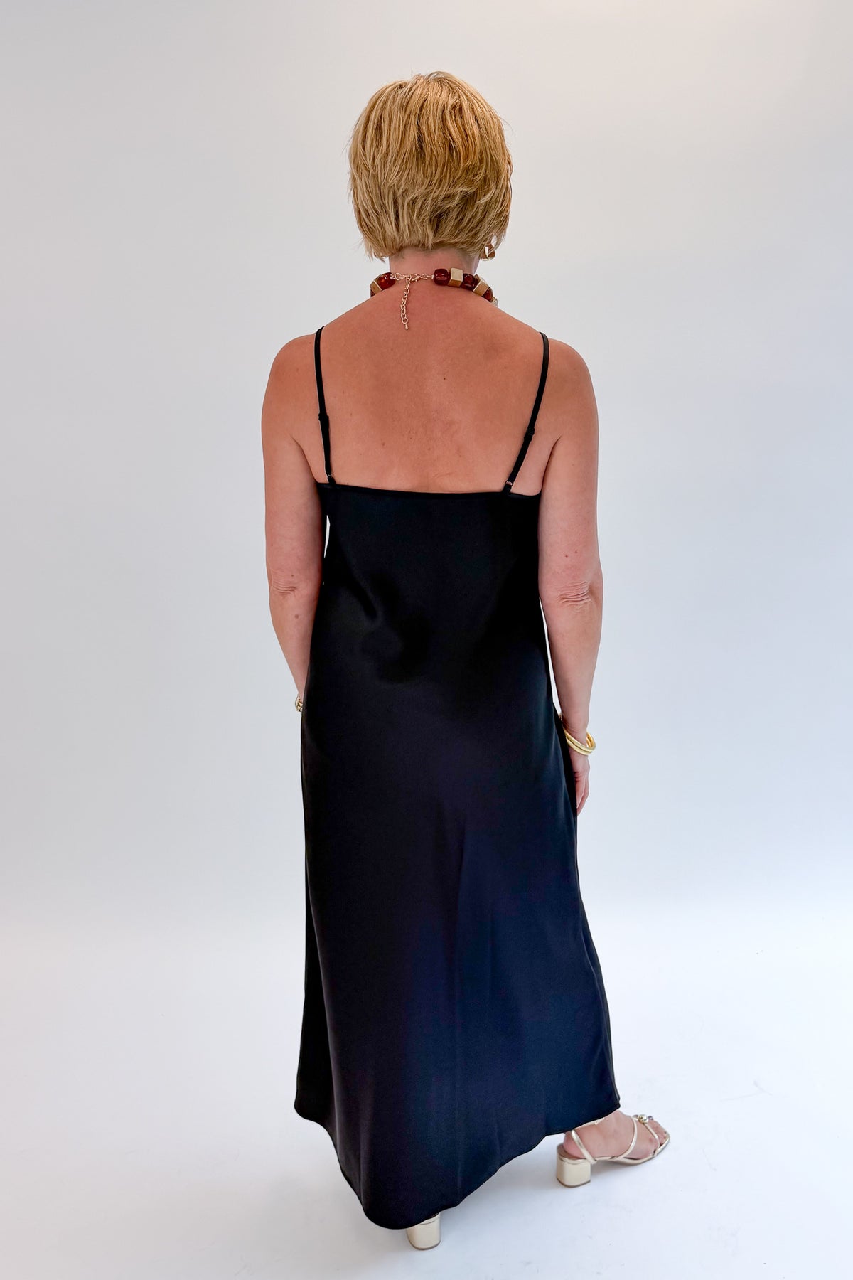 Back view of woman wearing Swagger Boutique’s Black Satin Dress with heels against white studio background.