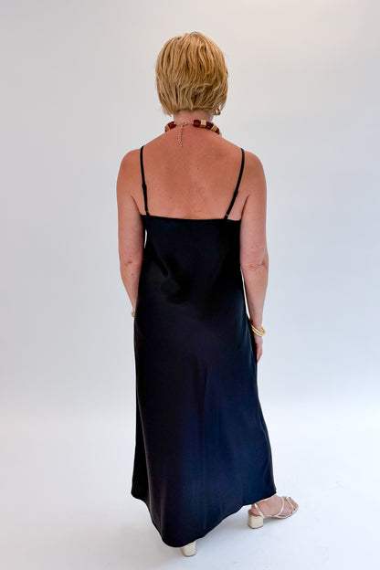 Back view of woman wearing Swagger Boutique’s Black Satin Dress with heels against white studio background.