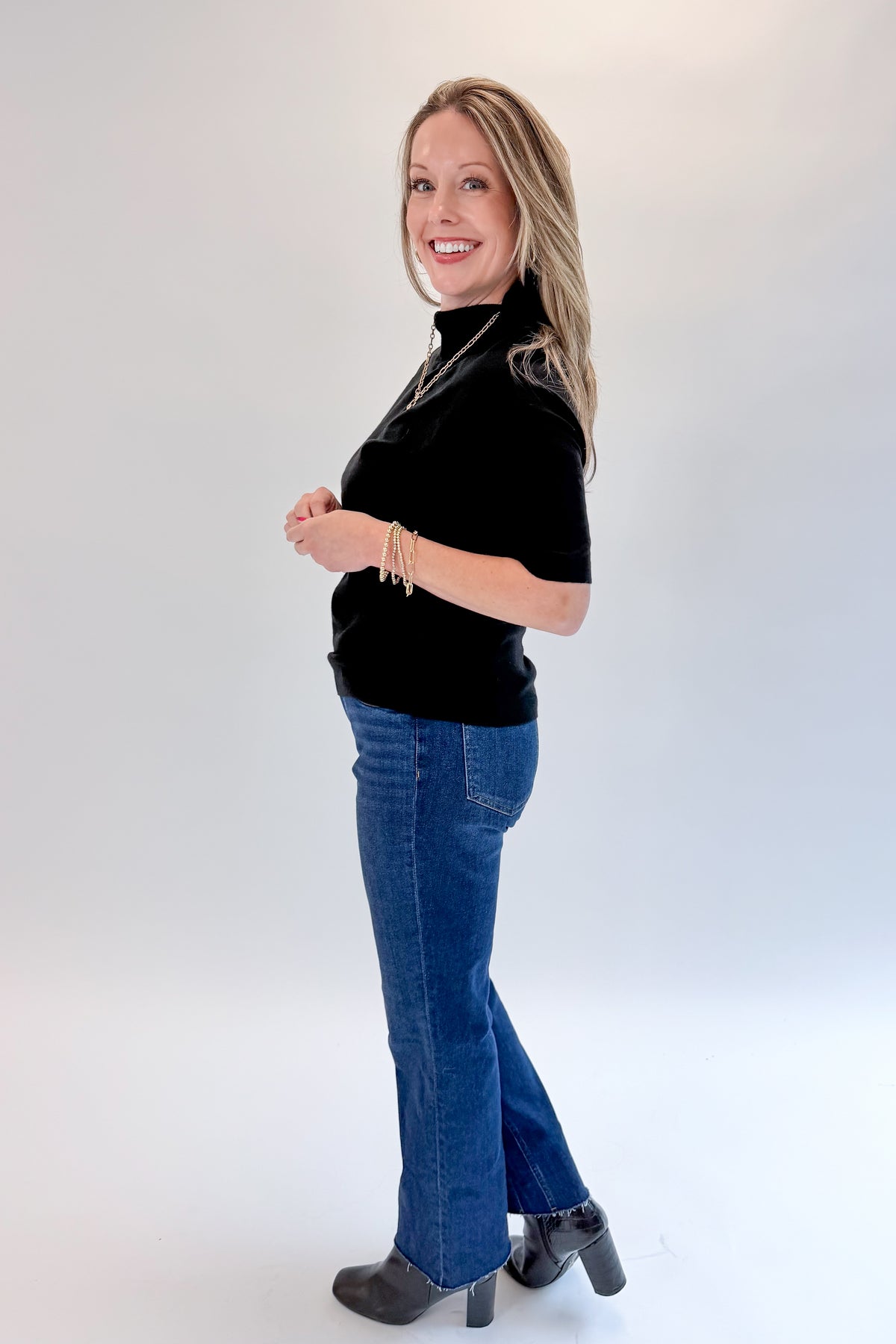 Side view of woman wearing Swagger Boutique’s Black Short Sleeve Turtleneck Sweater with jeans against white studio background.