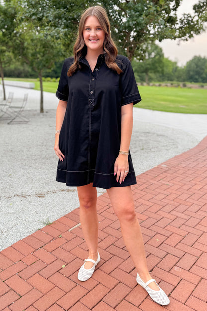 Front view of woman wearing Swagger Boutique’s Black Stitched Mini Dress with white flats in an outdoor scenery. 