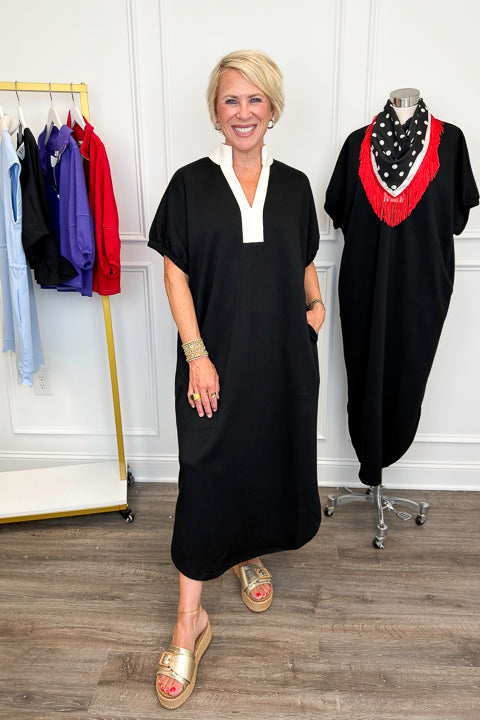 Front view of woman wearing Swagger Boutique’s Black With Cream Lining Midi Dress with sandals with clothing hanging in the background. 