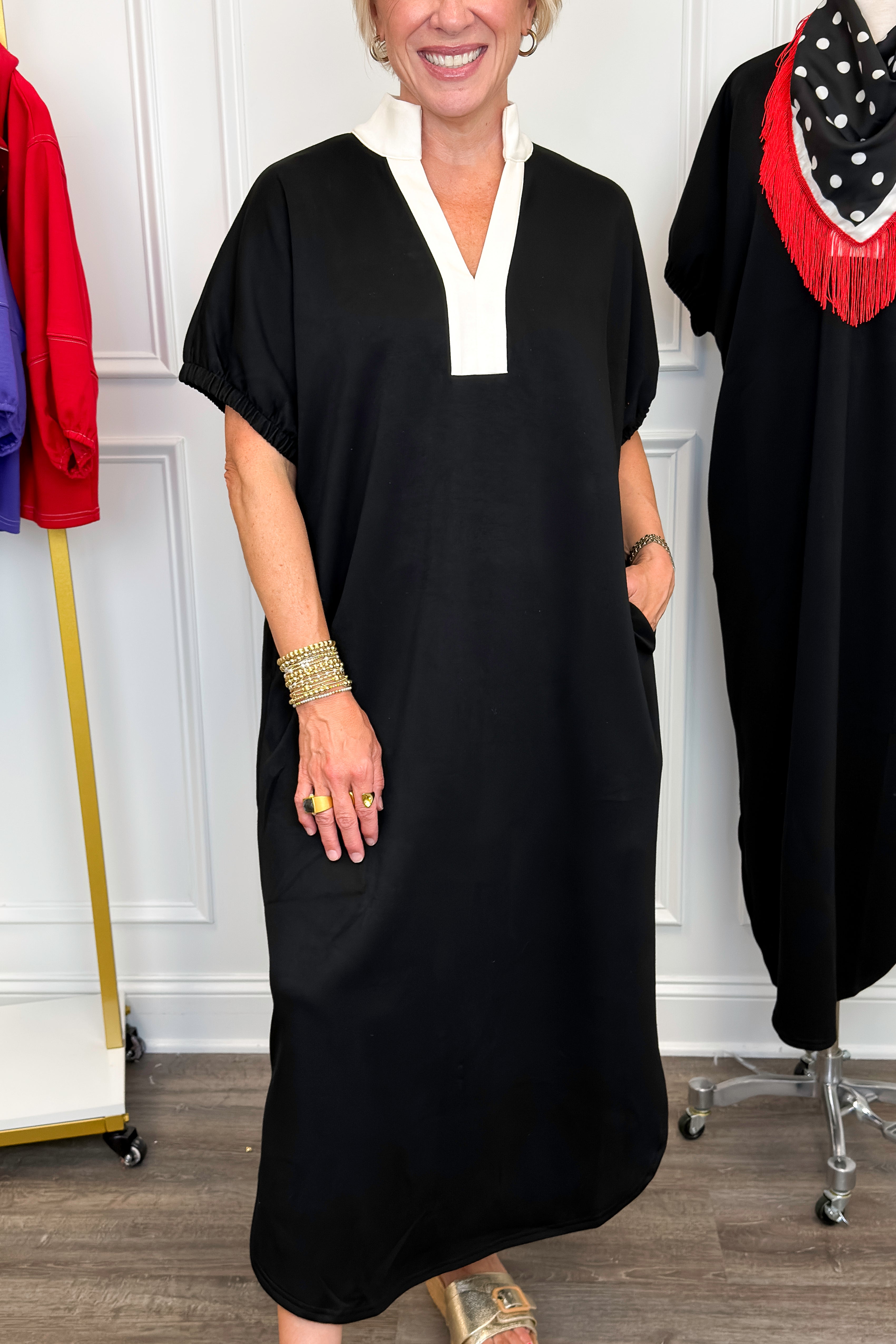 Front detail view of woman wearing Swagger Boutique’s Black With Cream Lining Midi Dress with sandals with clothing hanging in the background. 