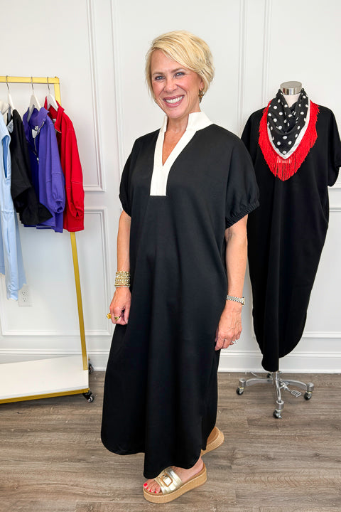 Side view of woman wearing Swagger Boutique’s Black With Cream Lining Midi Dress with sandals with clothing hanging in the background. 