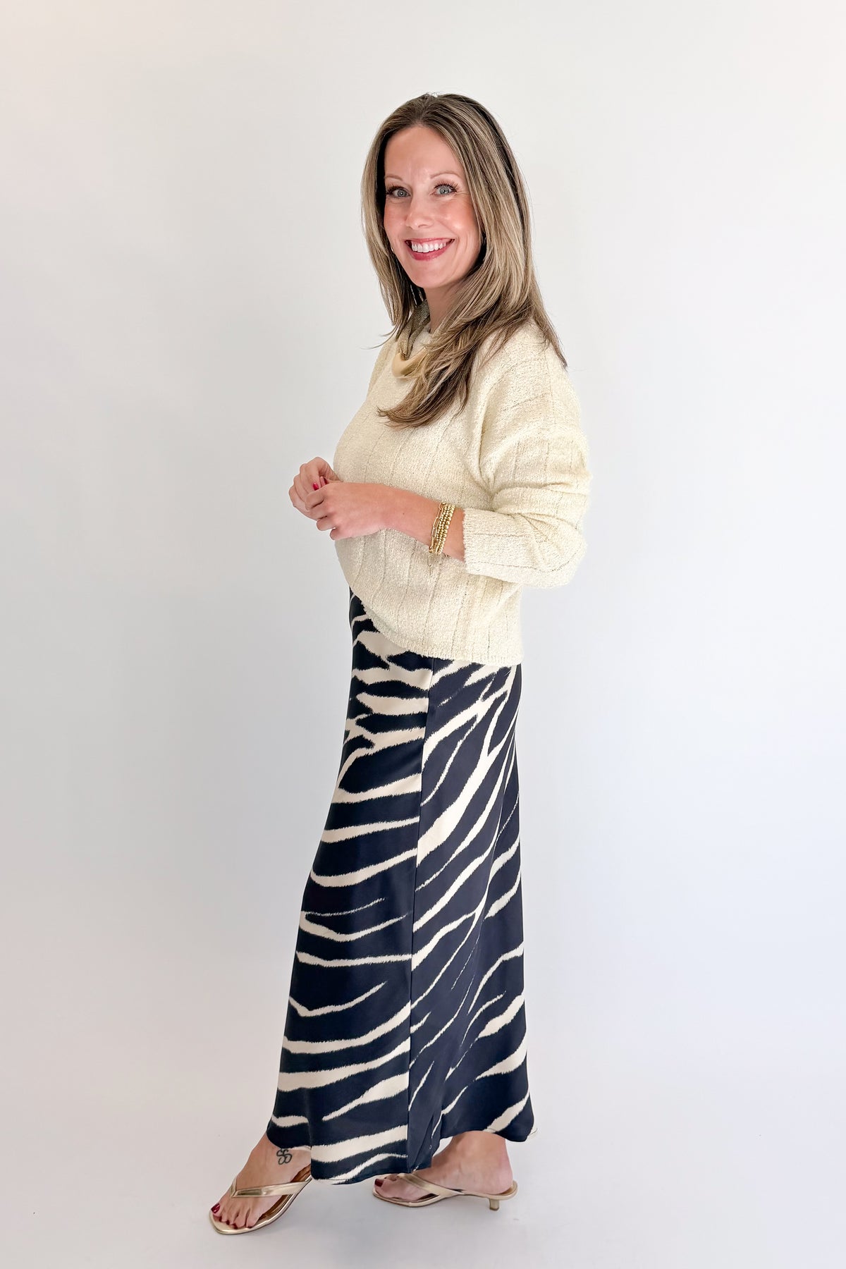 Side view of woman wearing Swagger Boutique’s Black Zebra Print Satin Skirt with Cream Ribbed Textured Sweater against white studio background.