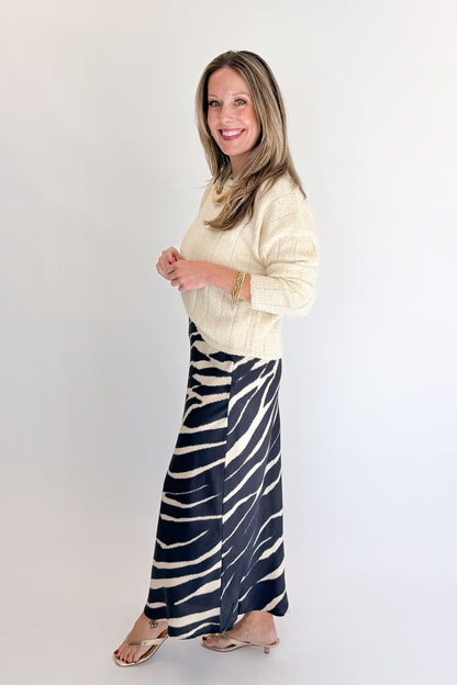 Side view of woman wearing Swagger Boutique’s Black Zebra Print Satin Skirt with Cream Ribbed Textured Sweater against white studio background.