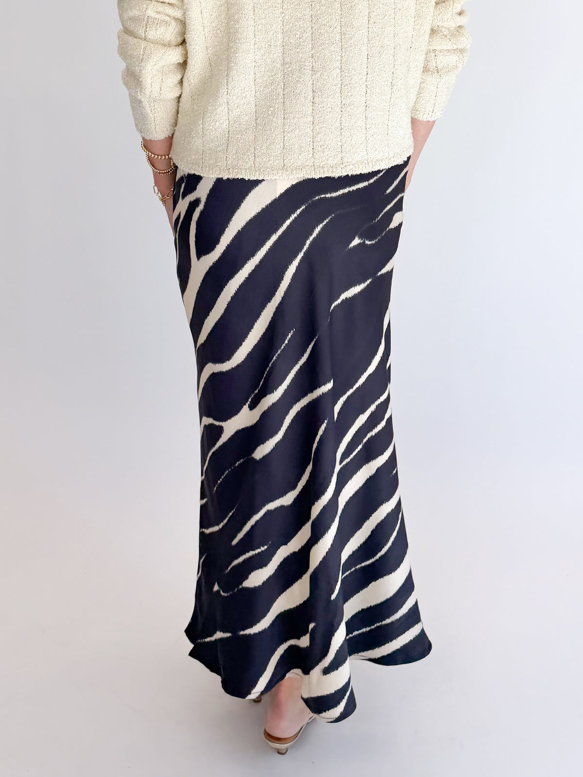 Back detail view of woman wearing Swagger Boutique’s Black Zebra Print Satin Skirt with Cream Ribbed Textured Sweater against white studio background.