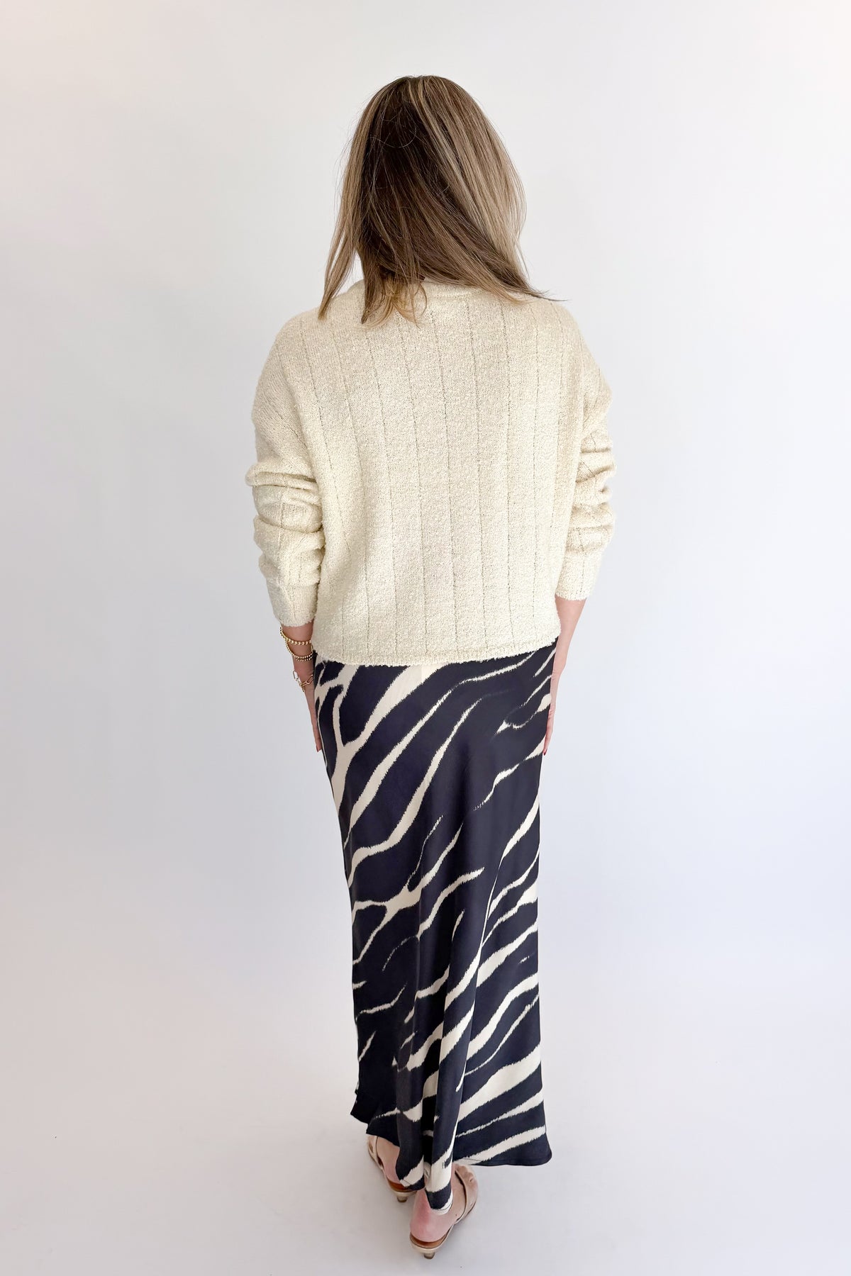 Back view of woman wearing Swagger Boutique’s Black Zebra Print Satin Skirt with Cream Ribbed Textured Sweater against white studio background.