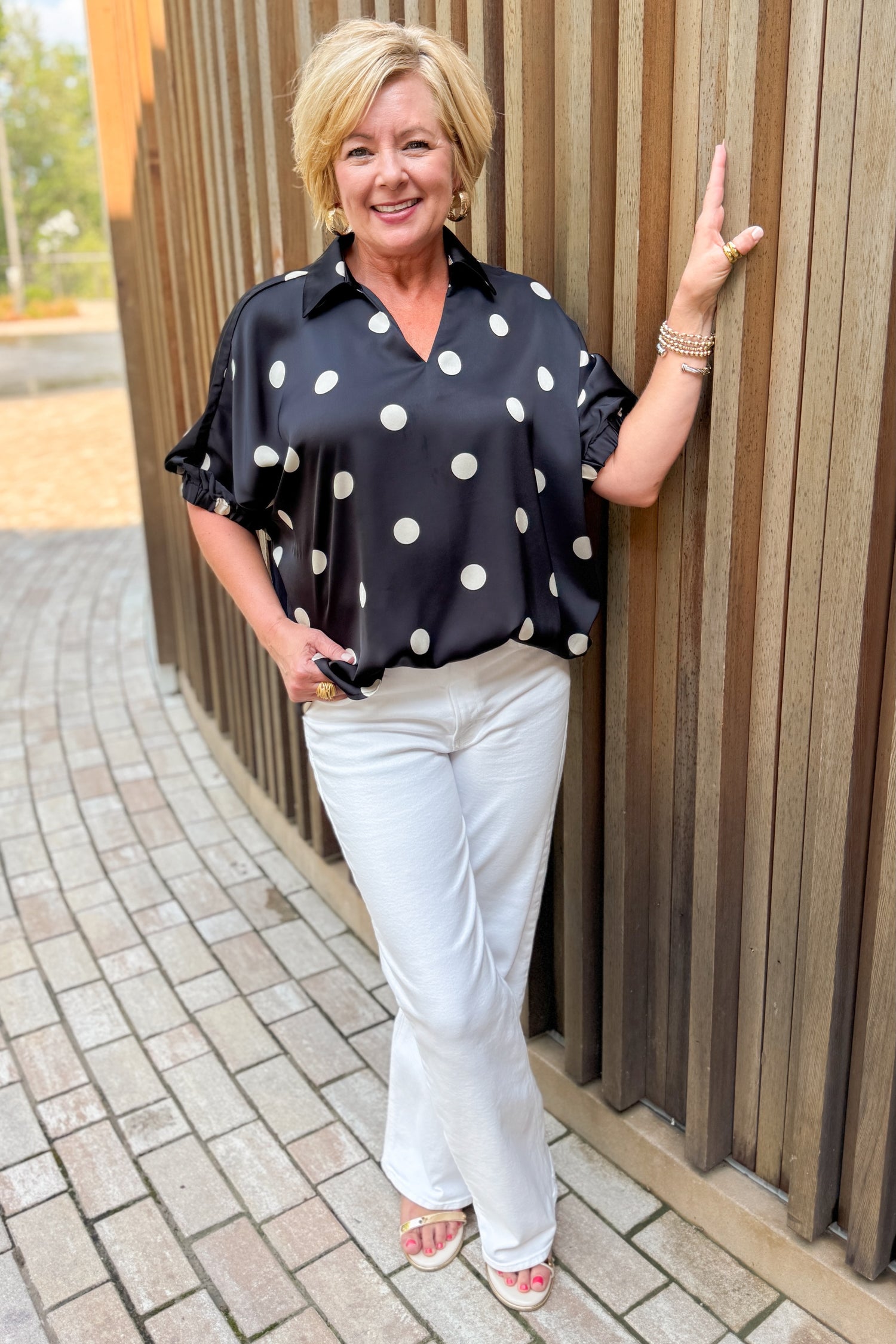 Front view of woman wearing Swagger Boutique’s Black &amp; Cream Polka Dot Satin Top with jeans standing outside on a brick path.