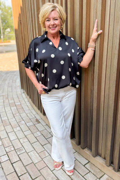Front view of woman wearing Swagger Boutique’s Black &amp; Cream Polka Dot Satin Top with jeans standing outside on a brick path.