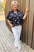 Front view of woman wearing Swagger Boutique’s Black & Cream Polka Dot Satin Top with jeans standing outside on a brick path.