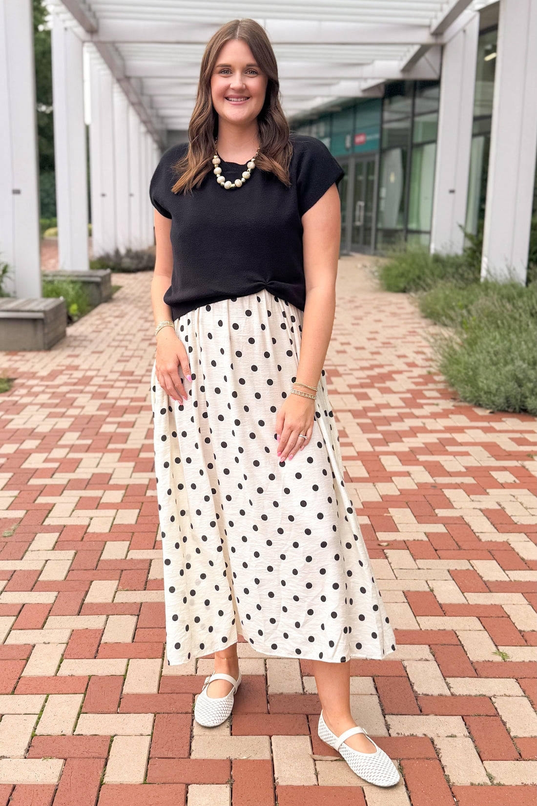 Front view of woman wearing Swagger Boutique’s Black &amp; Cream Polka Dot Layered Midi Dress by Entro outside by a red brick walkway.